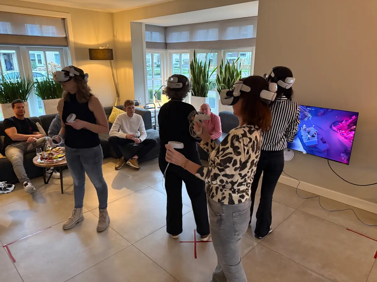 Spelers met VR-bril in een lounge, met het meekijkscherm aan de muur en toeschouwers op de bank